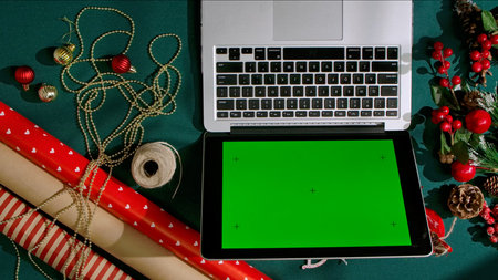 Top view laptop with chroma green screen on dark green festive background among wrapping paper, ornaments and twine, perfect mockup for holiday shopping, blogging, or festive design.の写真素材