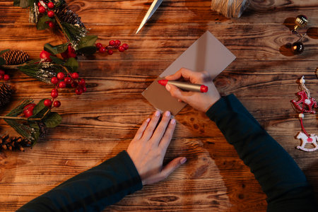 Top view of female hands writing a Christmas card with a red pen on a wooden table decorated with pinecones, red berries, and holiday ornaments. Warm festive atmosphere and cozy lighting.の写真素材