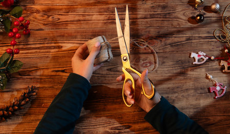 Close up top view of female hands using golden scissors to cut twine on a wooden table decorated with Christmas ornaments, red berries, and pinecones. Holiday crafting and DIY atmosphere.の写真素材