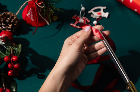 Female hands cutting shiny red ribbon with golden scissors on green surface surrounded by holiday decorations, twine, and ornaments in bright sunlight.の写真素材