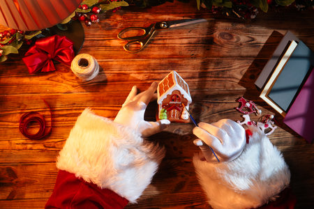 Top view of Santas gloved hands creating festive decorations with paint, ribbons, and scissors next to a wrapped gift on a rustic wooden table.の写真素材
