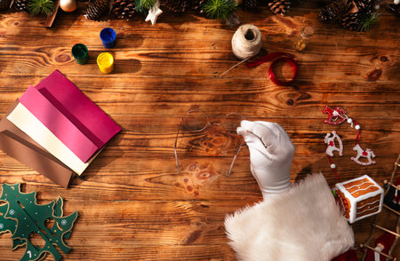 Santas gloved hands hold round gold rimmed glasses above a wooden table as he prepares to read letters and prepare gifts in his workshop.の写真素材
