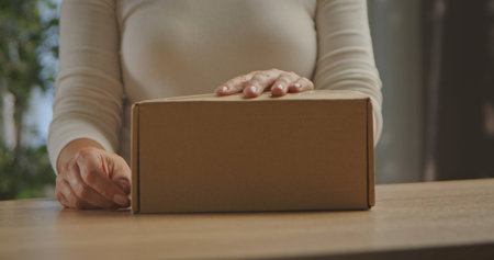 A woman in a white sweater placing her hands on a cardboard box on a wooden table, indoors.の写真素材