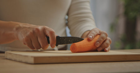 Close up of hands slicing a fresh carrot with a knife on a wooden cutting board in a kitchen...