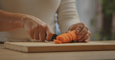 Close up of a person slicing a fresh carrot into thin pieces on a wooden cutting board.の写真素材