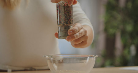 A person using a pepper grinder to add seasoning into a clear glass bowl, prepping ingredients for cooking.の写真素材