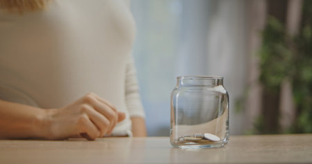A woman in a white shirt is sitting at a table, placing coins into a clear glass jar with a few coins inside.の写真素材