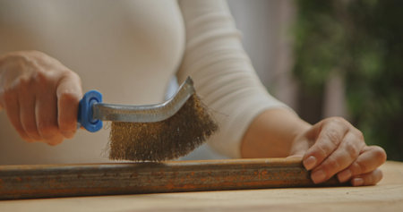 Close up of a woman using a wire brush to clean rust from a metal bar, focusing on the tool and technique.の写真素材