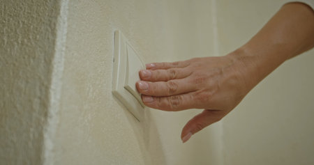 A woman hand is pressing a light switch mounted on a textured wall, demonstrating energy control.