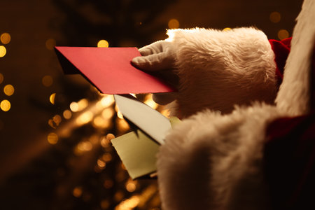 Santa Claus looks through envelopes with letters near a sparkling Christmas tree and warm lights in the background.の写真素材