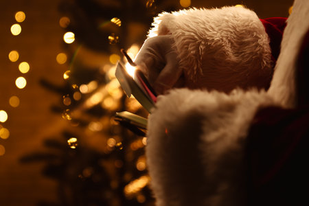 Santa Claus looks through envelopes with letters near a sparkling Christmas tree and warm lights in the background.の写真素材