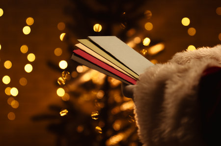 Santa Claus looks through envelopes with letters near a sparkling Christmas tree and warm lights in the background.の写真素材