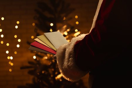 Santa Claus looks through envelopes with letters near a sparkling Christmas tree and warm lights in the background.の写真素材