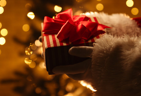 A festive scene featuring a Santa Claus figure holding a wrapped Christmas gift with a red ribbon in front of a beautifully lit Christmas tree.の写真素材