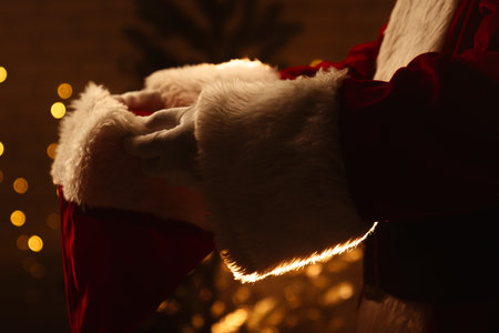 A close up image of Santa Claus holding his hat in front of a lit Christmas tree with warm lighting.の写真素材