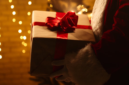 A festive scene featuring a Santa Claus figure holding a wrapped Christmas gift with a red ribbon in front of a beautifully lit Christmas tree.の写真素材