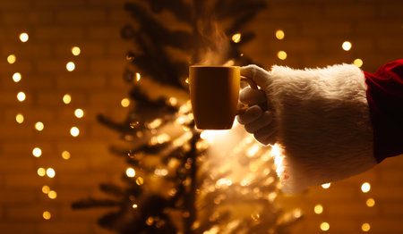 Close up of Santa Claus holding a hot drink near a Christmas tree, illuminated by warm festive lights. Festive mood on the eve of New Year Eve.の写真素材