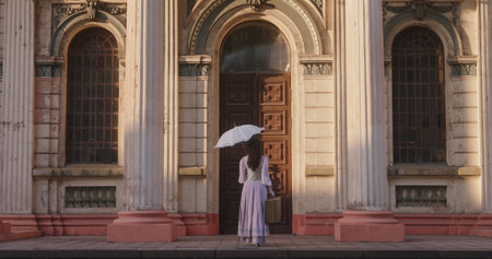 A woman in a historical dress holds a white umbrella and a suitcase, standing in front of majestic antique doors bathed in warm evening light, evoking a sense of anticipation and timeless beauty.の写真素材