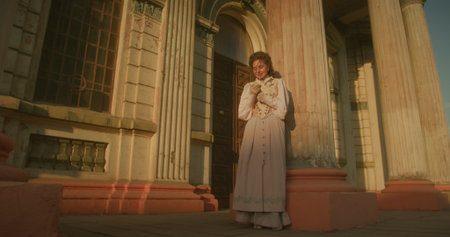 An elegant woman in a pastel historical dress stands next to a weathered column of an old building, with a dreamy, nostalgic expression on her face.の写真素材
