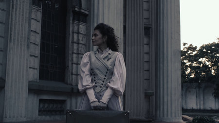 An elegant woman in historical attire stands at dusk near tall stone columns, holding a suitcase and...