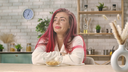 A woman with vibrant red hair sits pensively in a modern kitchen, wearing a cozy white robe with a bowl in front. Breakfast in the home kitchen, the morning ritual of eating.の写真素材