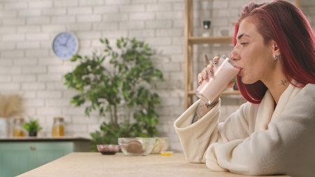 A young woman with red hair drinks a pink smoothie at a wooden table in a cozy, modern kitchen setting. Breakfast in the home kitchen, the morning ritual of eating.の写真素材