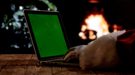 Santa Claus in a red suit and white gloves works on a laptop with a green screen chroma key in front of a fireplace and a Christmas tree. A warm festive atmosphere for digital or themed layouts.の写真素材