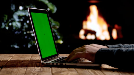 A man works on a laptop with a green screen chroma key background in front of a fireplace and a Christmas tree. A warm festive atmosphere for digital or themed layouts.の写真素材