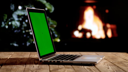 Laptop with a green screen chroma key screen against a glowing fireplace and Christmas tree. A warm, festive atmosphere for digital or themed layouts.の写真素材