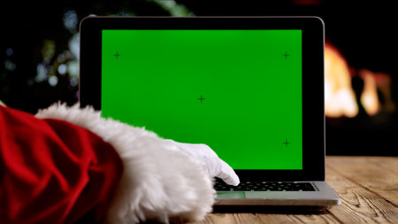 Santa Claus in a red suit and white gloves works on a laptop with a green screen chroma key in front of a fireplace and a Christmas tree. A warm festive atmosphere for digital or themed layouts.の写真素材