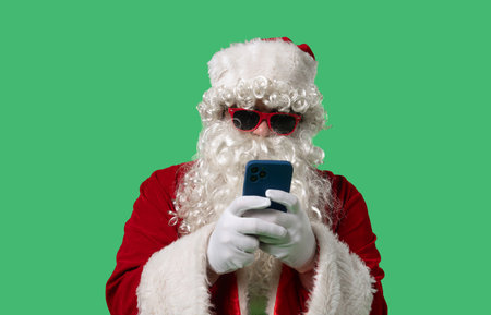 Santa Claus with a white curly beard, wearing a red suit and sunglasses, texting on a smartphone on...