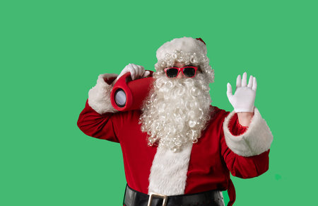 Santa Claus with a white curly beard, wearing a red suit, dances cheerfully with a portable speaker on a green background. Perfect for Christmas themed compositions.の写真素材
