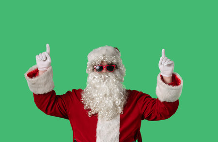Santa Claus with a white curly beard, wearing a red suit, points his fingers upward on a green background. Perfect for Christmas themed compositions, advertising, and holiday layouts.の写真素材