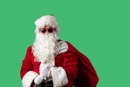 Santa Claus with a white curly beard, wearing a red suit and sunglasses, with a bag of gifts on a green background. Perfect for Christmas themed compositions, advertising, and holiday layouts.の写真素材