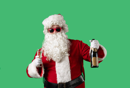 Santa Claus with a white curly beard, wearing a red suit, makes a congratulatory toast with a glass of champagne and a bottle in his hands against a green background.の写真素材