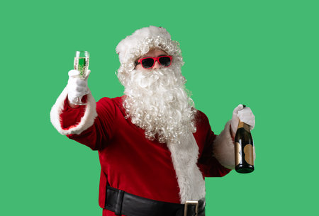 Santa Claus with a white curly beard, wearing a red suit and sunglasses, makes a toast with a glass of champagne and a bottle in his hands against a green background.の写真素材