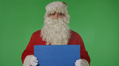 Santa Claus in his traditional red suit and white gloves holds a blank blue sign on a green chroma key background. Perfect for holiday advertising, greetings, or creative Christmas designs.の写真素材