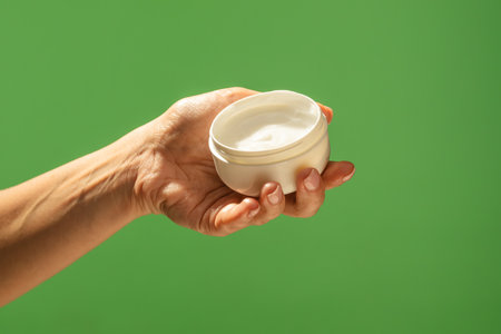A close up of a hand holding an open white cream jar against a vibrant green background. Beauty concept, skin care, and cream product presentation.の写真素材