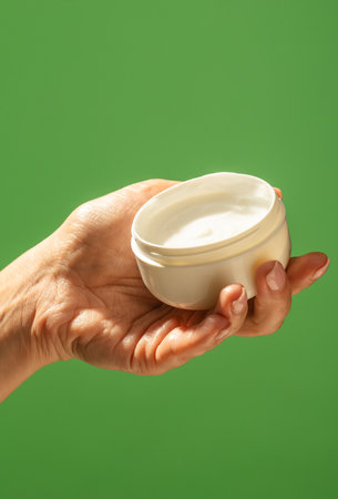 A close up of a hand holding an open white cream jar against a vibrant green background. Beauty...