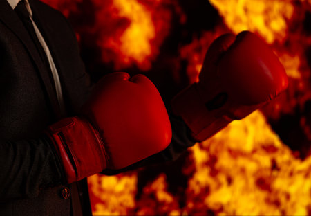 A determined businessman wearing a suit and red boxing gloves stands in front of a fiery background, symbolizing resilience and challenge.の写真素材