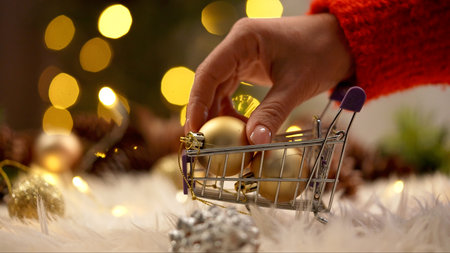 A hand picks up gold Christmas ornaments from a miniature shopping cart against a festive bokeh background.の写真素材