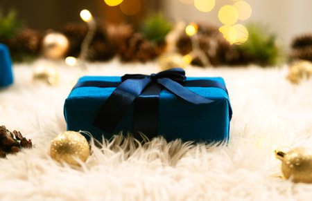 A blue gift box with a dark ribbon lies on a fluffy surface, surrounded by golden decorations and the blurred lights of a Christmas tree.の写真素材