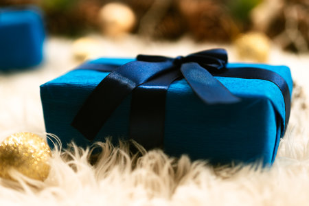 A blue gift box with a dark ribbon lies on a fluffy surface, surrounded by golden decorations and the blurred lights of a Christmas tree.の写真素材