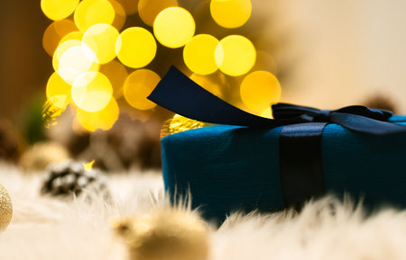 A blue gift box with a dark ribbon lies on a fluffy surface, surrounded by golden decorations and the blurred lights of a Christmas tree.の写真素材