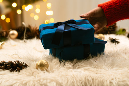 A cozy holiday scene with a hand holding a blue gift on a soft surface, surrounded by festive lights and decor.の写真素材