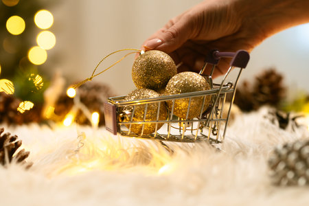 A hand picks up gold Christmas ornaments from a miniature shopping cart against a festive bokeh background.の写真素材
