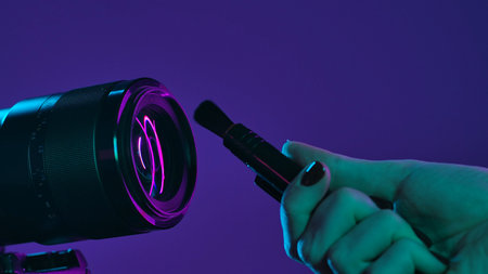 A hand carefully holds a brush near a camera lens, illuminated by vibrant neon purple and green lighting. Concept of cleaning and caring for photographic equipment.の写真素材