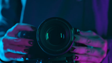 A close up shot of a camera lens held by a person, illuminated by vibrant purple and blue neon lights.の写真素材
