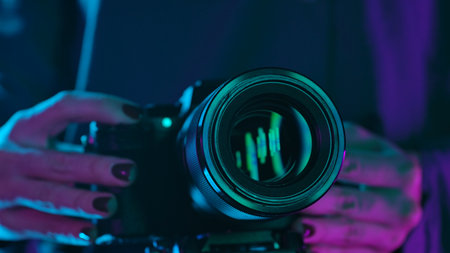 A close up shot of a camera lens held by a person, illuminated by vibrant purple and blue neon...