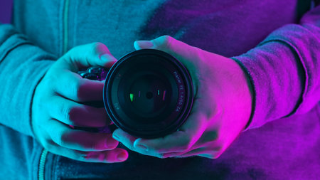 Close up of hands holding a camera lens with vibrant pink and blue neon lighting in a dark setting.の写真素材